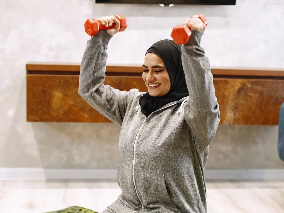 Smiling woman in hijab and hoodie lifting light dumbbells on a mat